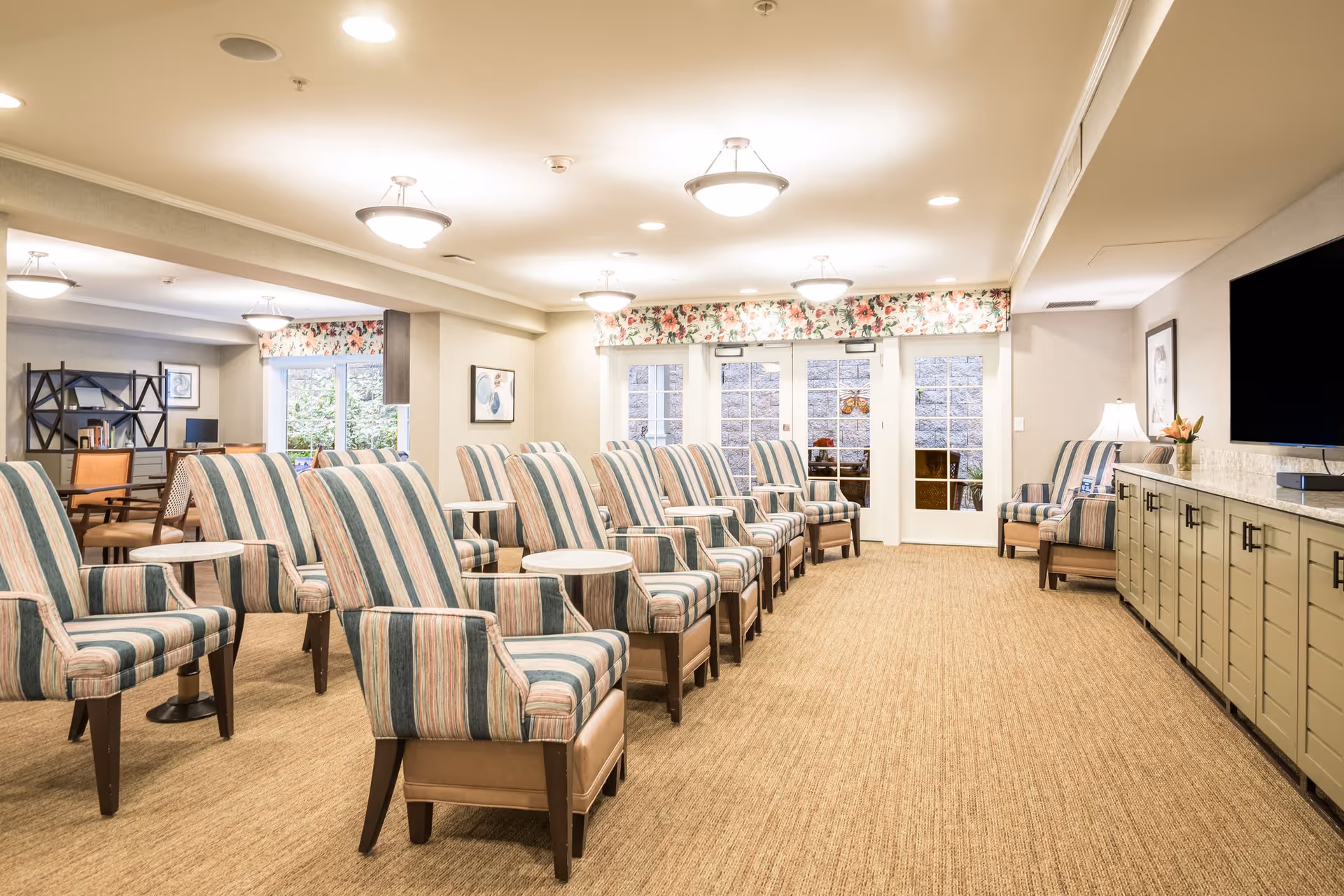 A well-lit common area with multiple striped armchairs arranged in rows facing a large flat-screen TV mounted on the wall. The room has beige carpeting, floral valances above glass doors, and additional seating and a desk area in the background.