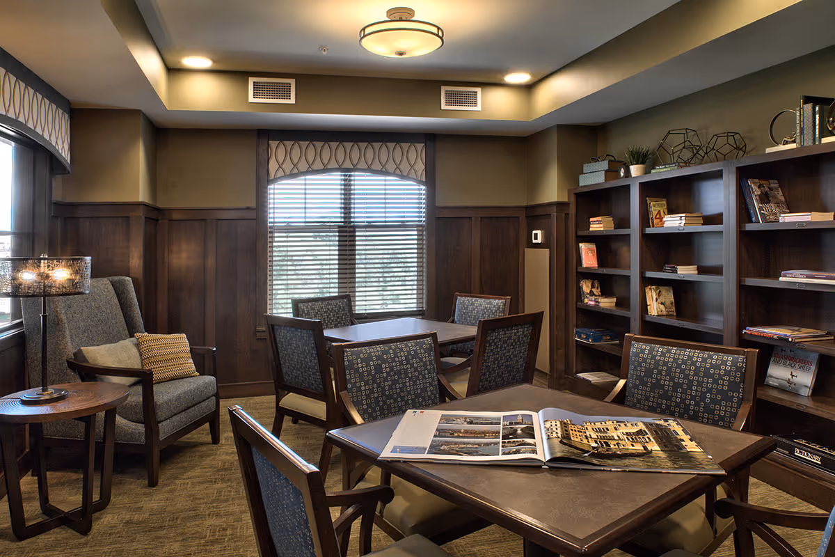 A cozy interior room with wooden paneling and shelves filled with books. There are multiple tables with patterned chairs around them, and a comfortable armchair with a side table and lamp. A large window with blinds and a decorative valance lets in natural light.