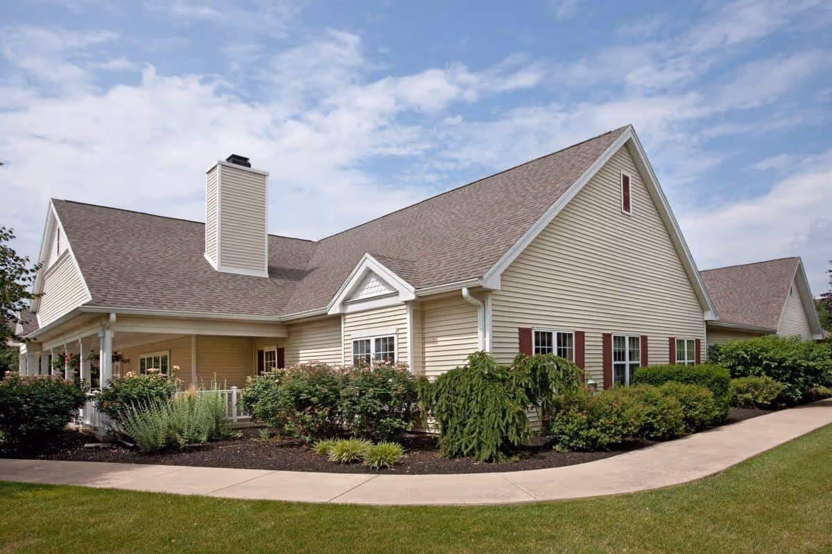 Exterior view of a single-story senior living facility building with beige siding, a chimney, multiple windows with red shutters, surrounded by well-maintained bushes and a curved concrete walkway under a partly cloudy sky.