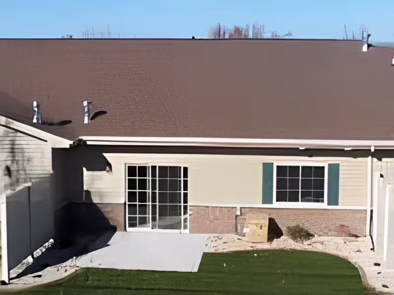 Exterior wall of a single-story building with a brown roof, sliding glass door, window with green shutters, and a small patio area with turf.