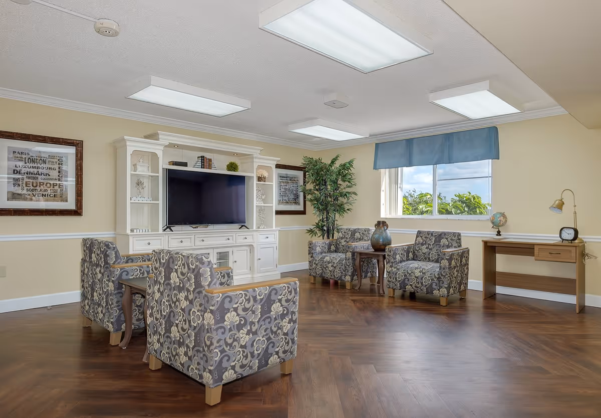 A cozy living room area with four patterned armchairs arranged around a small wooden table with a decorative vase. There is a large white entertainment center with a flat-screen TV and shelves holding books and decor. The room has wood flooring, beige walls with white trim, a window with a blue valance, a potted plant, framed artwork on the walls, and a wooden desk with a globe, clock, and lamp.