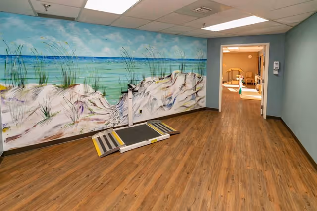 Interior room with wood-look flooring, a large beach mural on one wall, a wheelchair transfer ramp on the floor, and a doorway into another room.