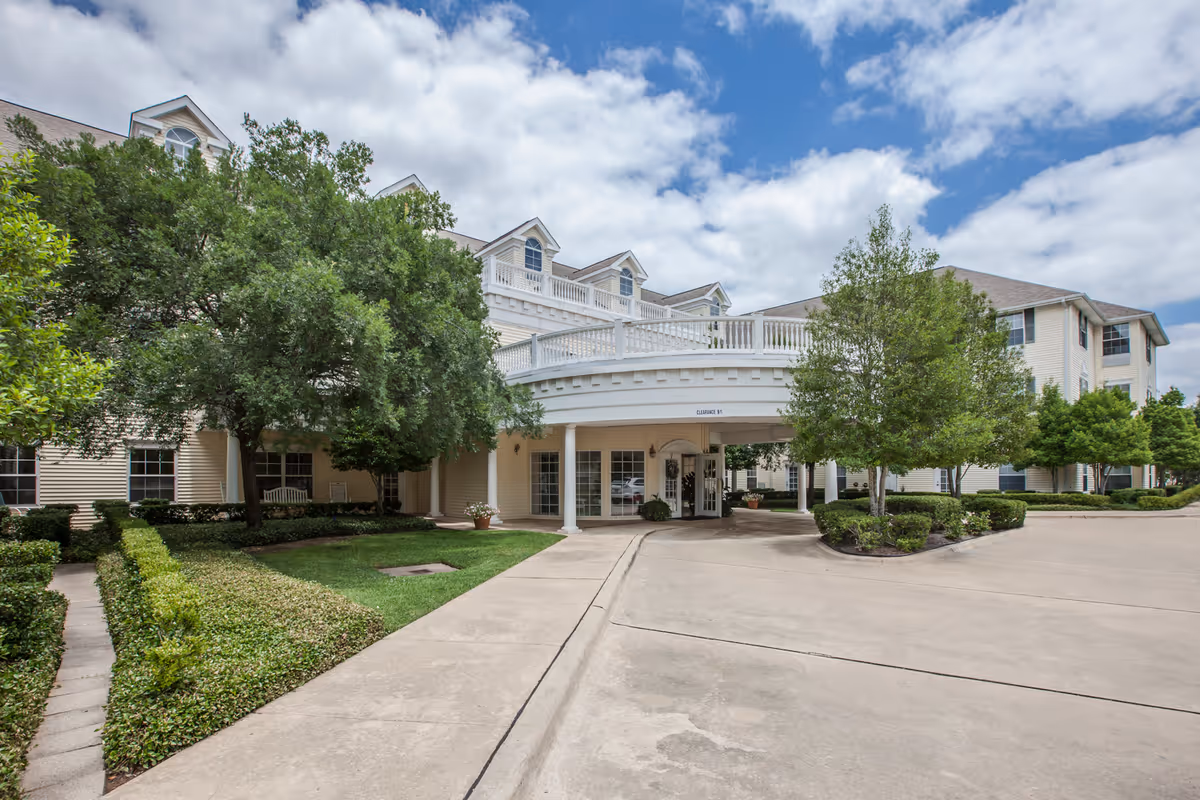 Front entrance of Bloom at Bossier senior living building with a porte-cochere, driveway and landscaped trees under a partly cloudy sky.