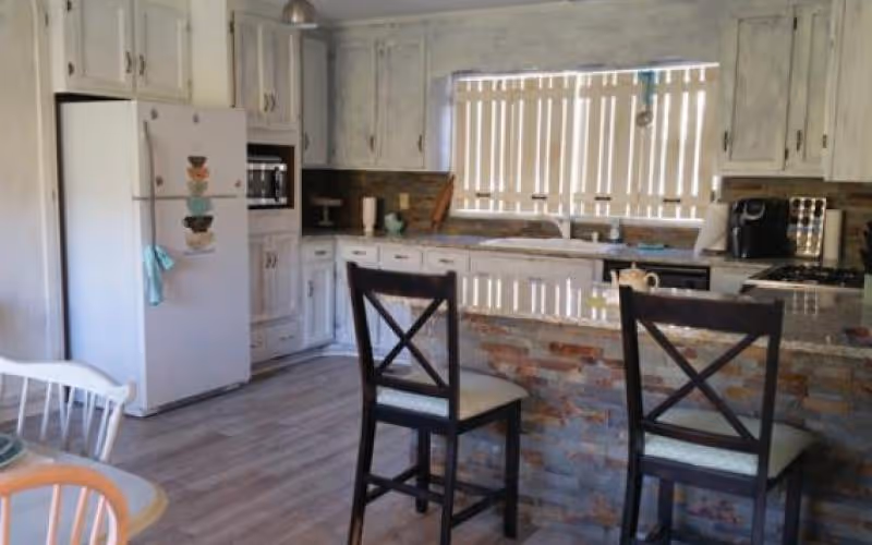 A kitchen with white cabinets, a white refrigerator, a built-in microwave, and a stove. There is a kitchen island with a stone facade and a polished countertop, with two dark wooden chairs with cushioned seats. A window with vertical blinds is above the sink, and a coffee maker is on the counter.