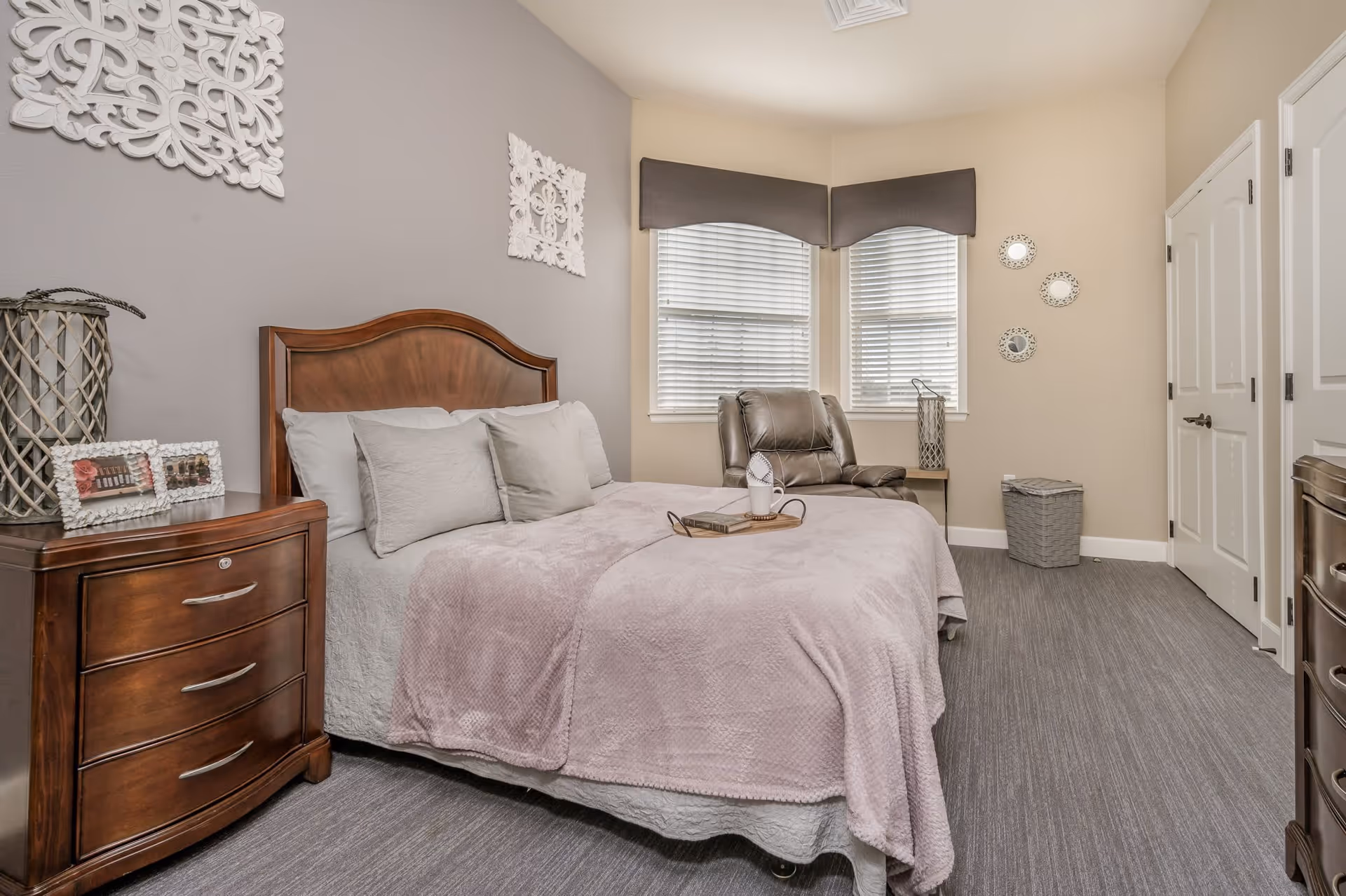A cozy bedroom with a wooden headboard bed covered in light gray and lavender bedding. There are three pillows on the bed and a tray with a book and a decorative item. To the left of the bed is a wooden nightstand with two framed photos and a decorative lantern. The walls are painted in two tones, gray and beige, with white decorative wall hangings. In the corner near two windows with white blinds and dark valances, there is a brown leather recliner chair. On the right side, there are two white doors and a gray laundry basket on the floor.