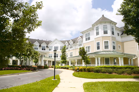 Exterior view of a large, multi-story senior living facility building with light yellow siding, white trim, and multiple windows. The building is surrounded by green lawns, trees, and a paved walkway under a partly cloudy sky.