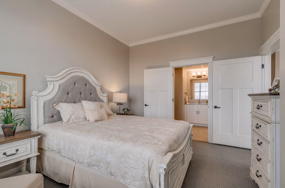 A bedroom with a large bed featuring an ornate white and gray tufted headboard, beige patterned bedding, and multiple pillows. There is a nightstand with a lamp and a plant on the left side of the bed, a dresser on the right side of the room, and an open door leading to a bathroom with a vanity and mirror visible.