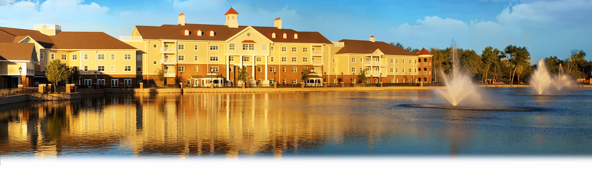 A large, multi-story senior living facility named Live Oak with a red roof and yellow exterior walls, situated beside a calm lake with two water fountains spraying water into the air. The building and fountains are reflected in the lake, and there are trees and a clear blue sky in the background.