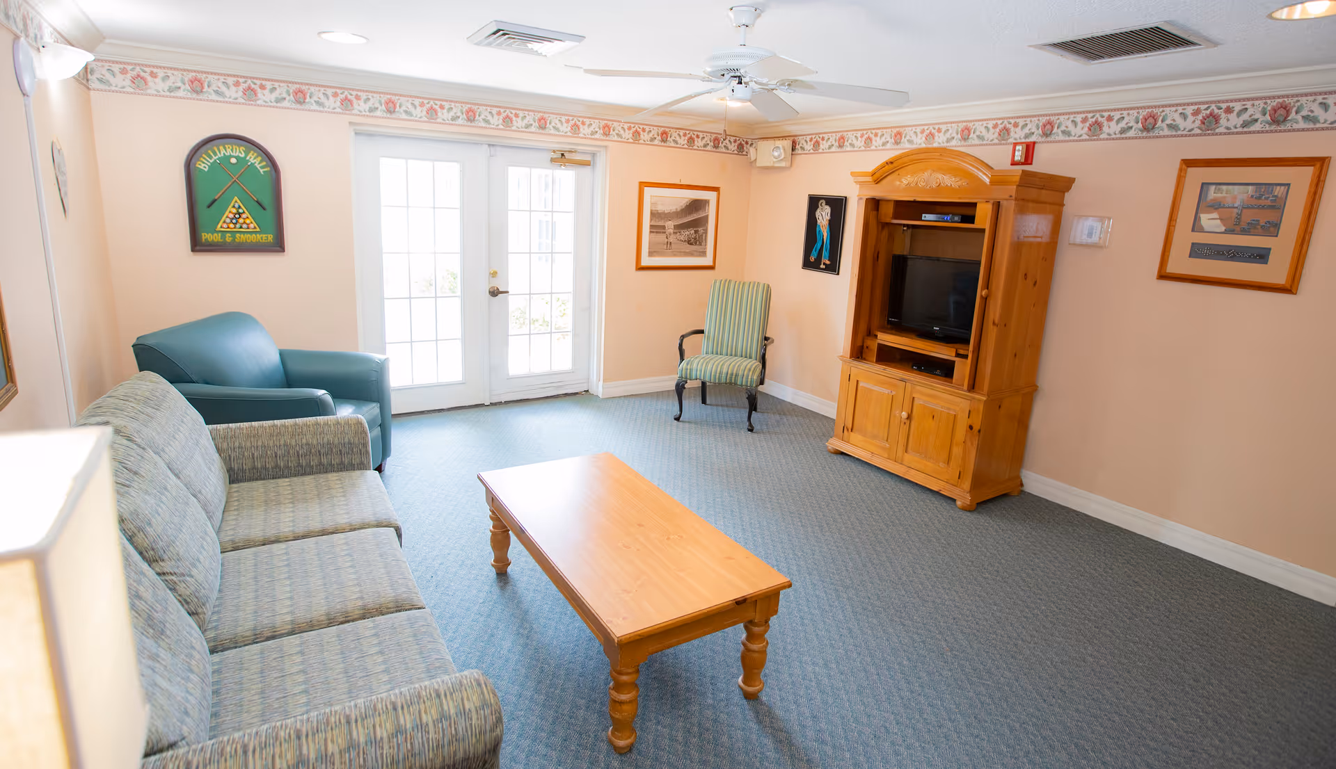 A cozy living room with a patterned sofa, a green armchair, a wooden coffee table, and a wooden entertainment center with a TV. The room has light peach walls with a floral border near the ceiling, framed pictures on the walls, a ceiling fan, and double glass doors letting in natural light.