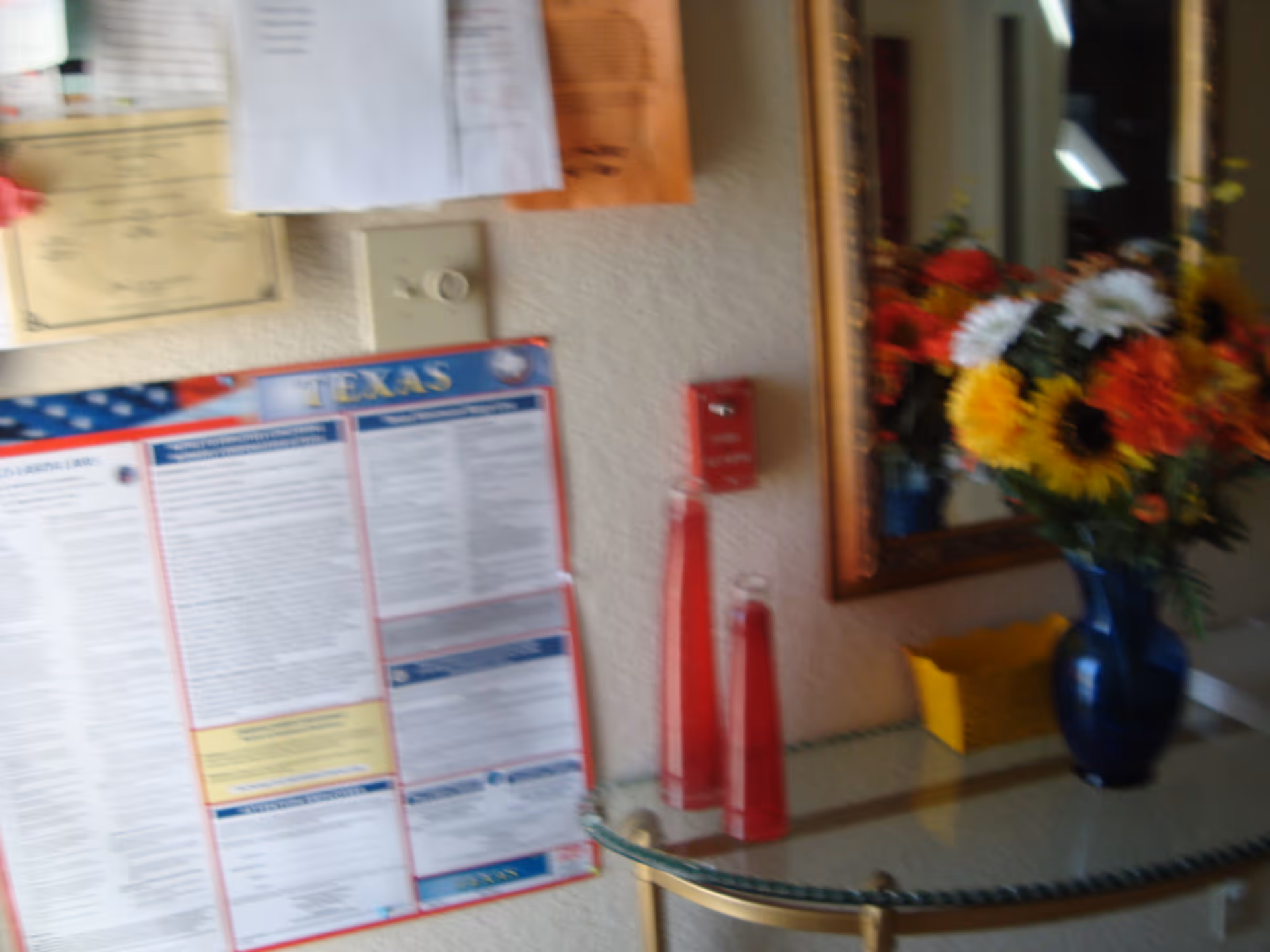 Glass-topped console table with a blue vase of flowers beneath a mirror and a Texas bulletin board with notices on the wall.