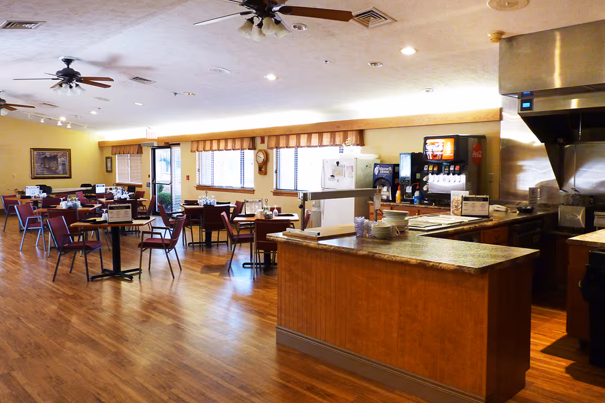 Interior view of a dining room in a senior living facility with wooden floors, multiple tables and chairs arranged for dining, ceiling fans, and a serving counter with plates and a beverage dispenser.