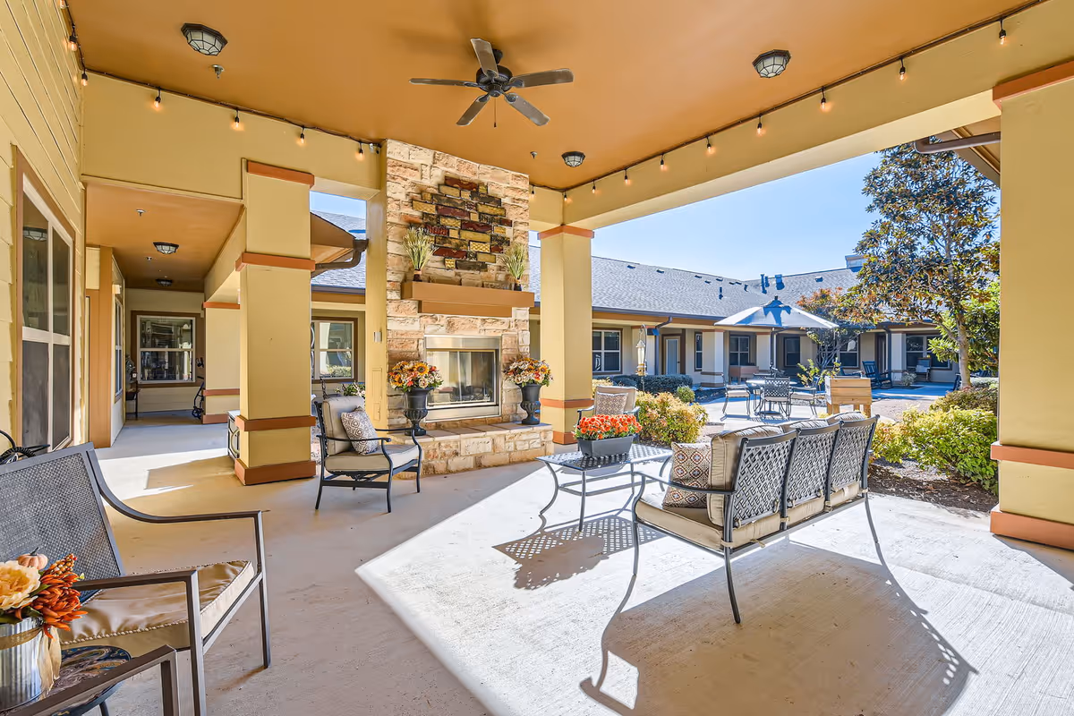 Covered outdoor patio area at Orchard Park of Kyle Assisted & Senior Living featuring cushioned metal chairs and a loveseat around a stone fireplace. The patio overlooks a courtyard with additional seating, greenery, and a tree under a clear blue sky.