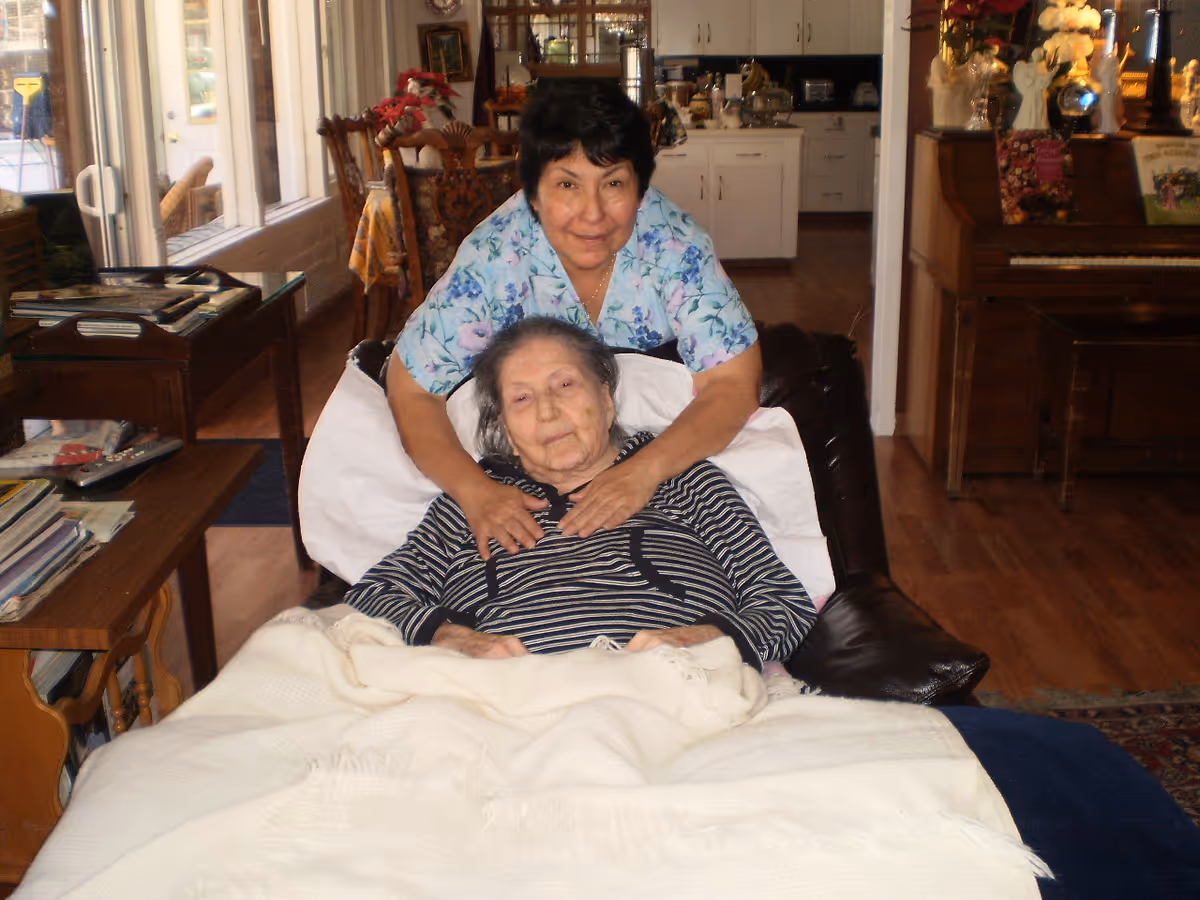 An elderly woman lying in a reclined chair covered with a white blanket, with a caregiver standing behind her, gently placing hands on her shoulders. The setting appears to be a cozy living room with wooden floors, a piano, and a dining area in the background.