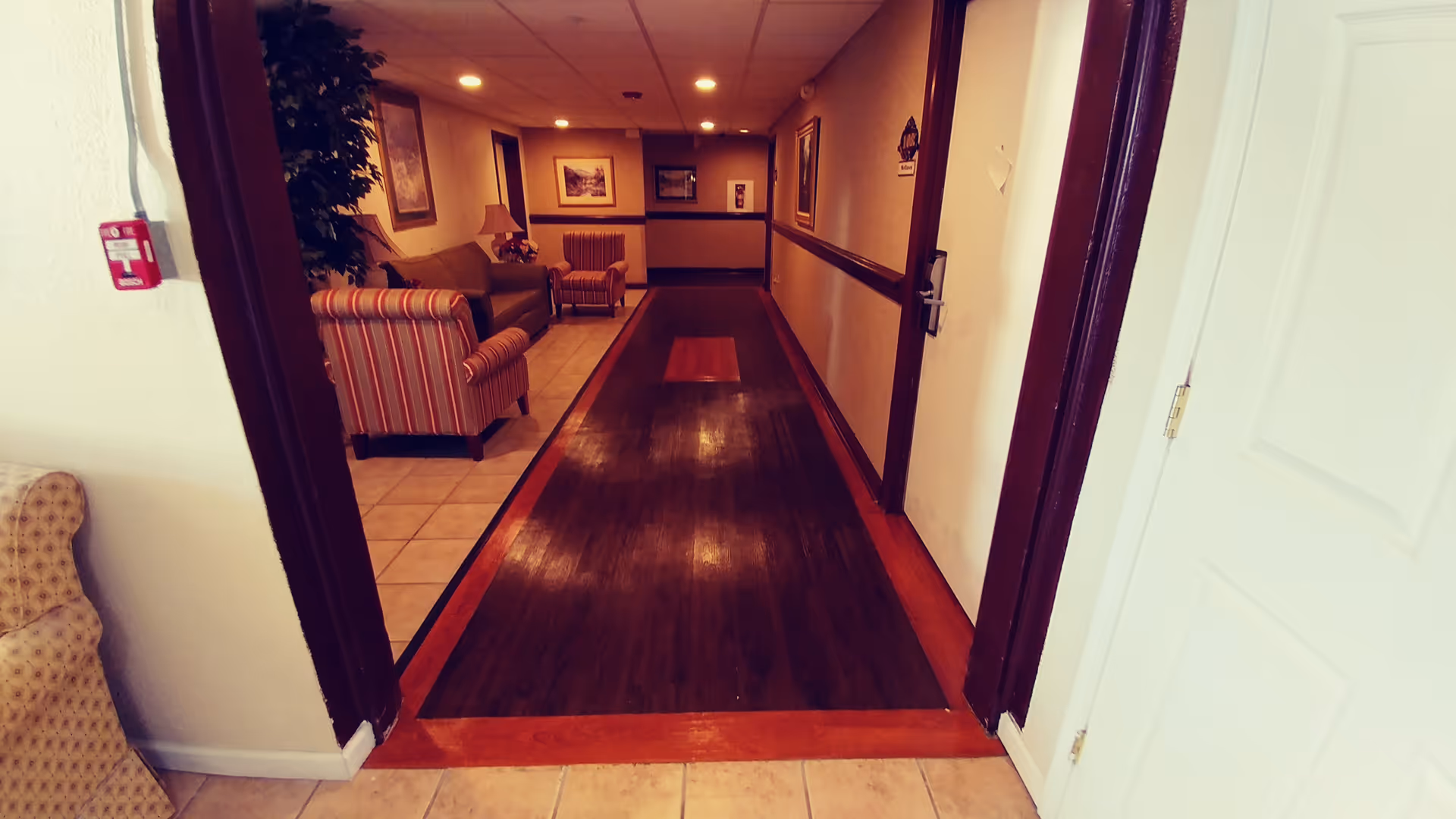 View down a hallway in a senior living facility with a seating area on the left featuring striped and solid armchairs and framed artwork on the walls. The hallway has a dark wooden floor bordered by lighter tiles and beige walls with a dark chair rail.