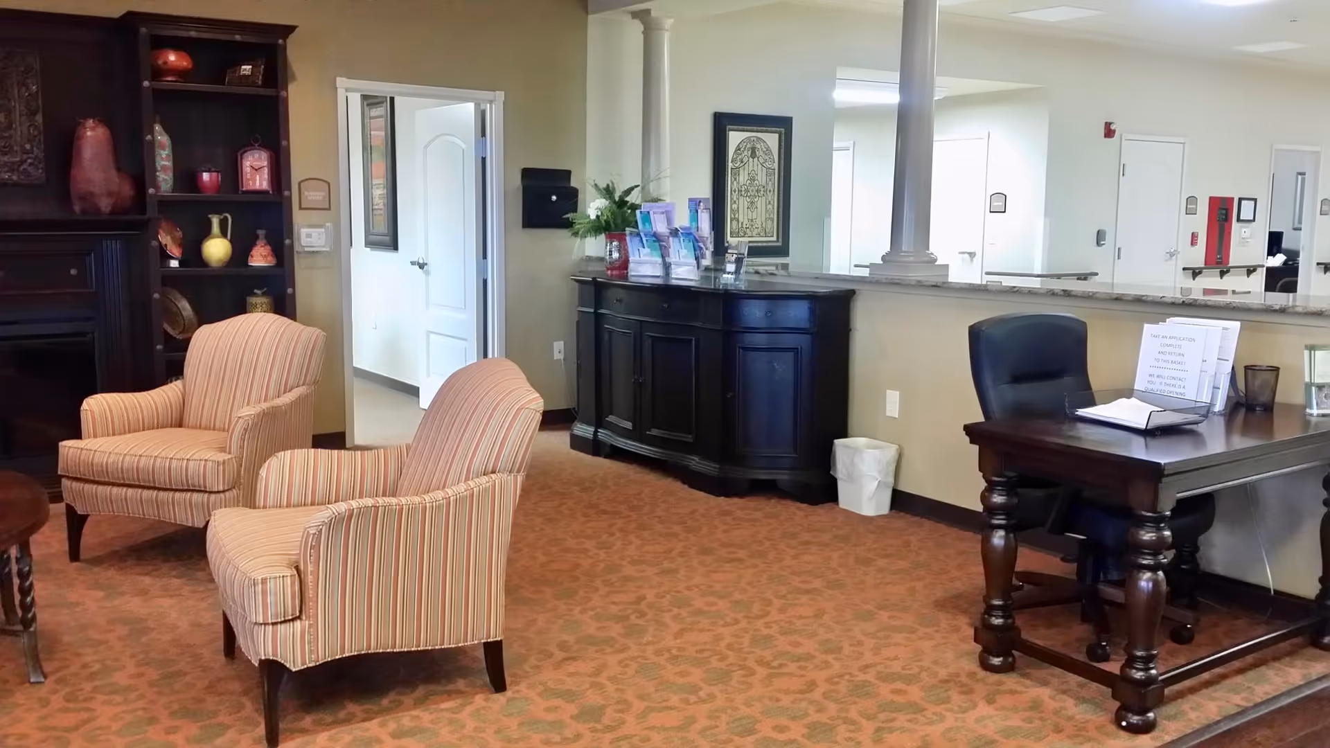 A cozy interior room with two striped upholstered armchairs on a patterned carpet. There is a dark wooden bookshelf with decorative items on the left, a black sideboard with brochures and a plant in the center, and a wooden desk with a black office chair on the right. The walls are light-colored, and there is an open doorway leading to another room.