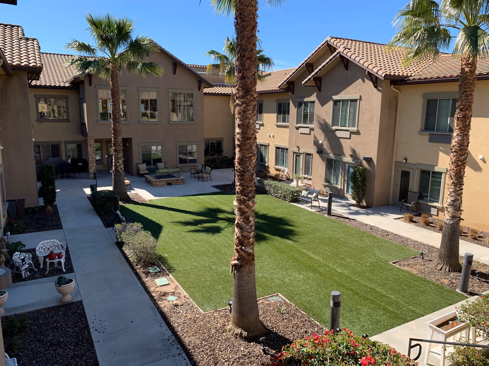 Outdoor courtyard area of a senior living facility with a well-maintained green lawn, several palm trees, and surrounding two-story beige buildings with tiled roofs. There are outdoor seating areas with chairs and tables along the pathways and near the building entrances under a clear blue sky.