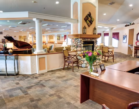 A spacious and well-lit senior living facility common area featuring a stone fireplace with a wooden mantle, several chairs arranged around it, a grand piano on the left side, and a wooden reception desk with a vase of flowers in the foreground. The room has carpeted floors, columns, and large windows with curtains allowing natural light to enter.