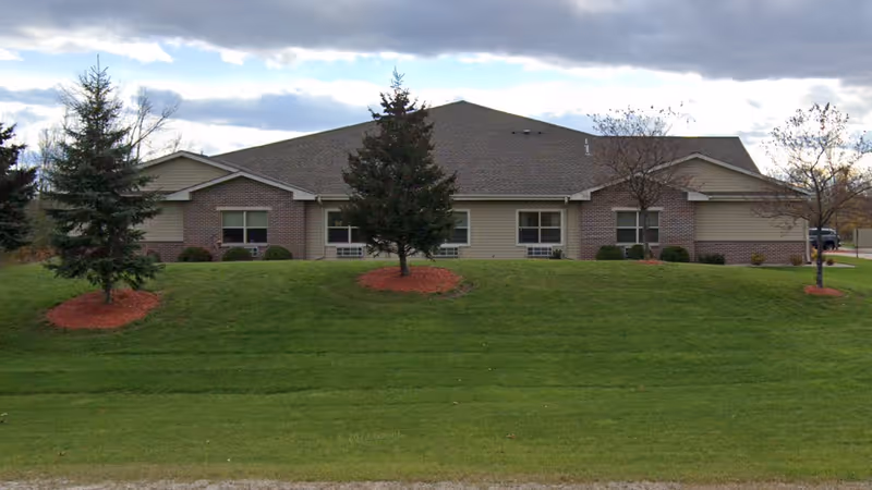 Front exterior view of a low, single-story assisted living facility with green lawn and trees.