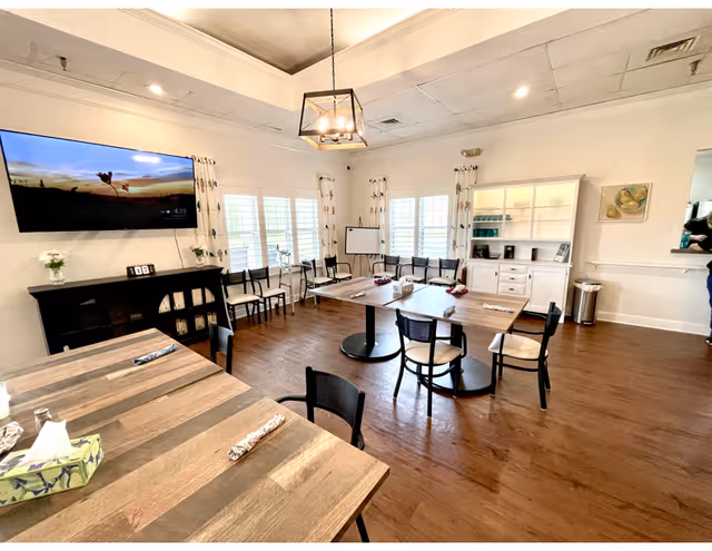 A bright and spacious common area with wooden floors and white walls. The room features several tables with chairs arranged around them, a large flat-screen TV mounted on the wall, a white cabinet with shelves holding cups and a coffee maker, and windows with white shutters and patterned curtains. A modern chandelier hangs from the ceiling, and there is a whiteboard in the corner.