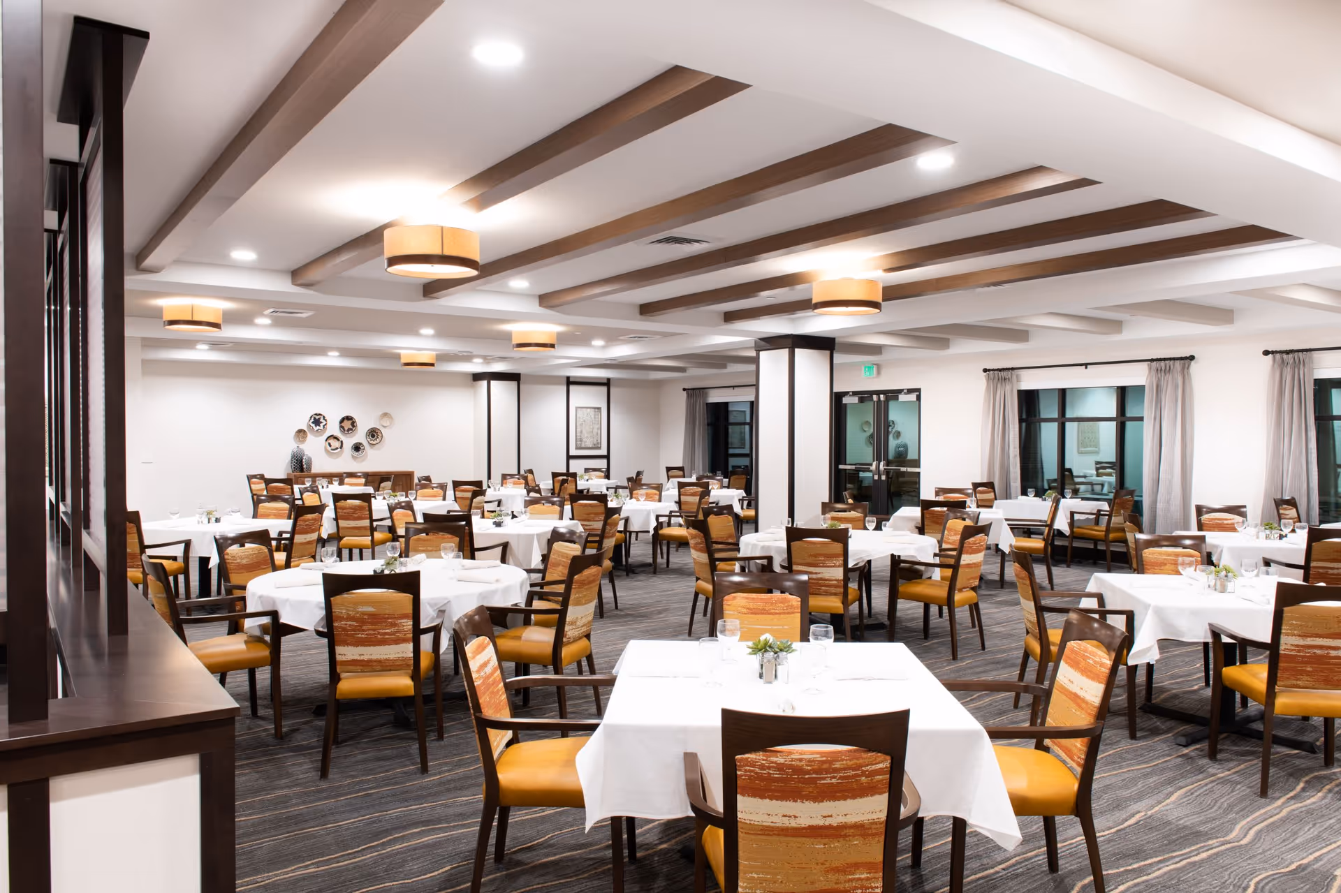 A spacious dining room with multiple tables covered in white tablecloths, each surrounded by wooden chairs with orange and brown patterned cushions. The room features a modern ceiling with recessed lighting and circular hanging light fixtures. Large windows with curtains and decorative wall plates are visible in the background.