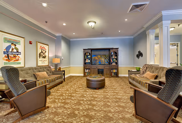 A cozy living room with two striped sofas facing each other and two armchairs. A wooden entertainment center with a TV and bookshelves is against the far wall. The room has beige patterned carpet, light blue walls with white trim, and framed vintage movie posters on one wall. A round ottoman is centered on the carpet, and a table lamp is on a side table next to one sofa.