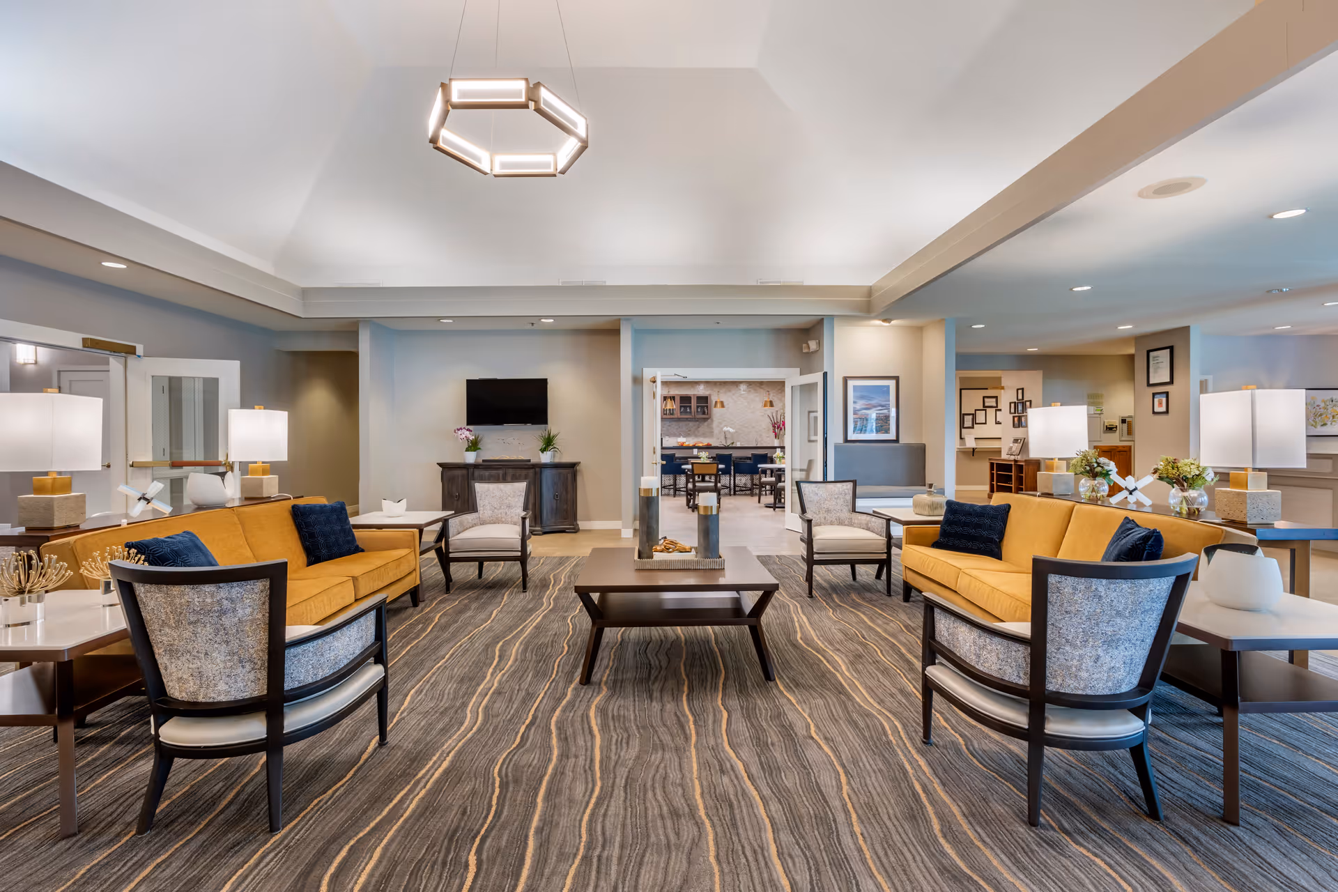 A spacious and well-lit living room area in a senior living facility featuring two mustard yellow sofas, four patterned armchairs, multiple side tables with lamps, a central coffee table with decorative items, and a modern hexagonal ceiling light fixture. In the background, there is a TV mounted on a cabinet and an open doorway leading to a dining area with tables and chairs.