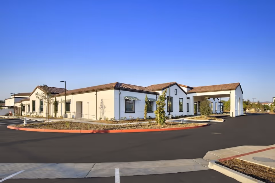 Front exterior of a single-story stucco building with a tile roof, porte-cochere and landscaped circular driveway under a clear blue sky.