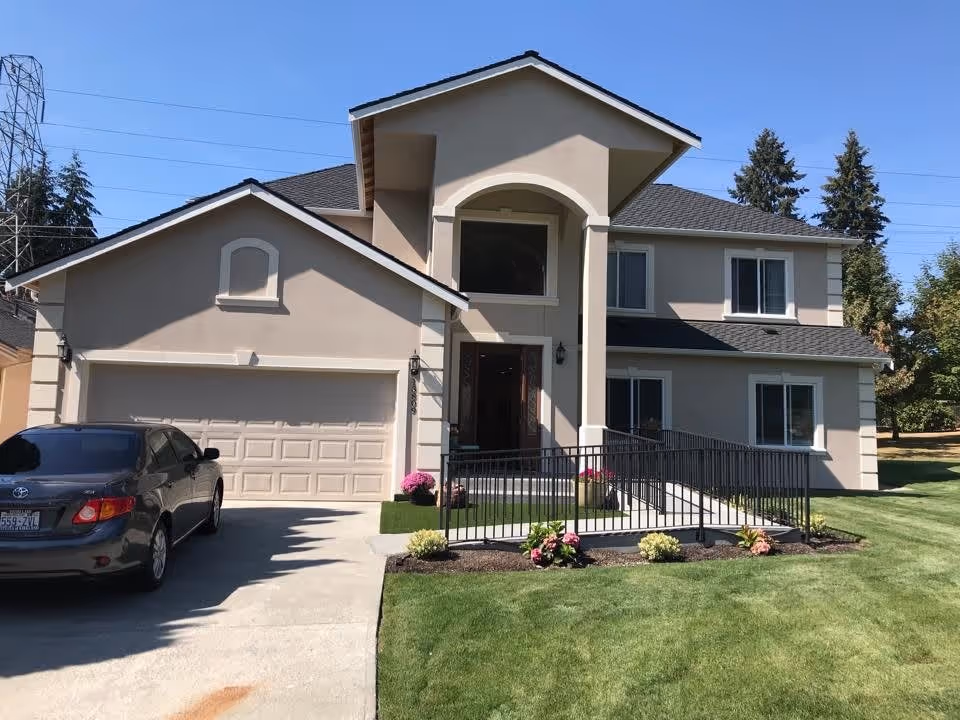 Front exterior view of a two-story beige house with a dark gray roof, a garage, a black car parked in the driveway, a wheelchair ramp leading to the front door, and a well-maintained lawn with small flower beds.