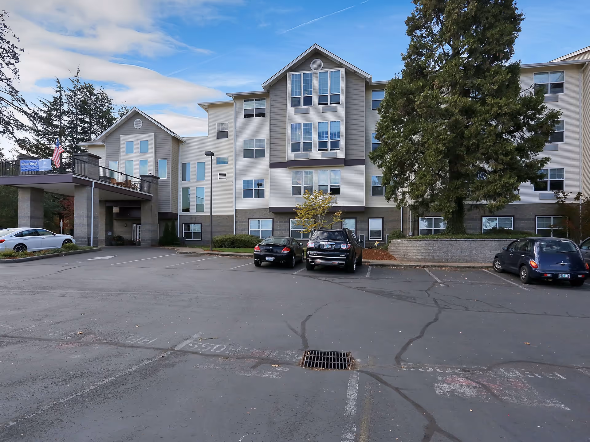Exterior view of a multi-story senior living facility building with several windows, a covered entrance, a large tree, and a parking lot with a few parked cars under a partly cloudy sky.