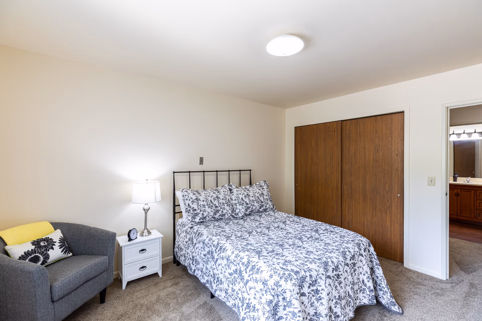 A bedroom with a bed covered in a white and blue floral bedspread, a gray armchair with a yellow blanket and a decorative pillow, a white nightstand with a lamp and a small clock, a wooden closet with sliding doors, and an open doorway leading to a bathroom.