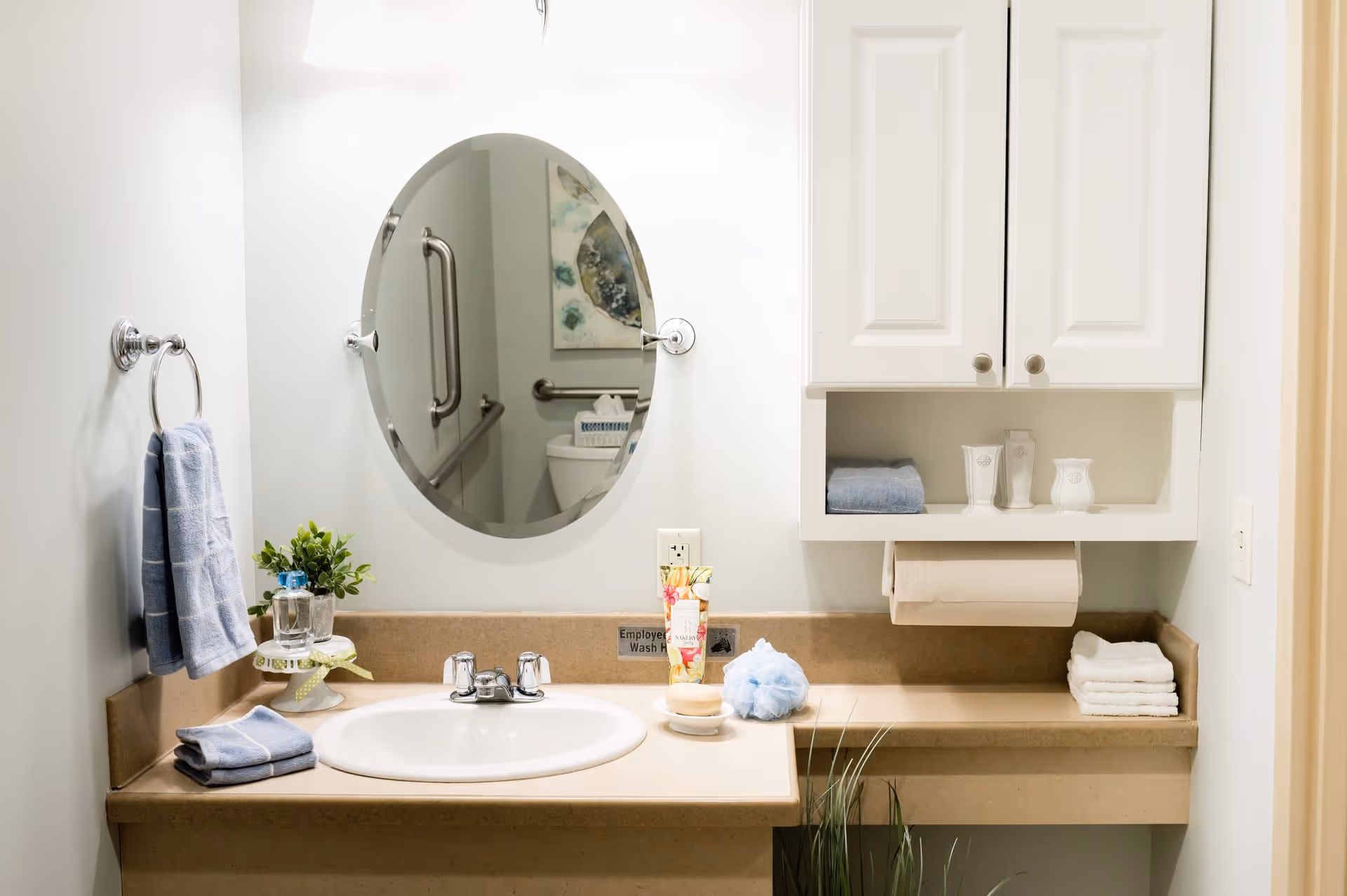 Well-lit bathroom sink area with an oval mirror, countertop toiletries, folded towels, and a wall-mounted cabinet.