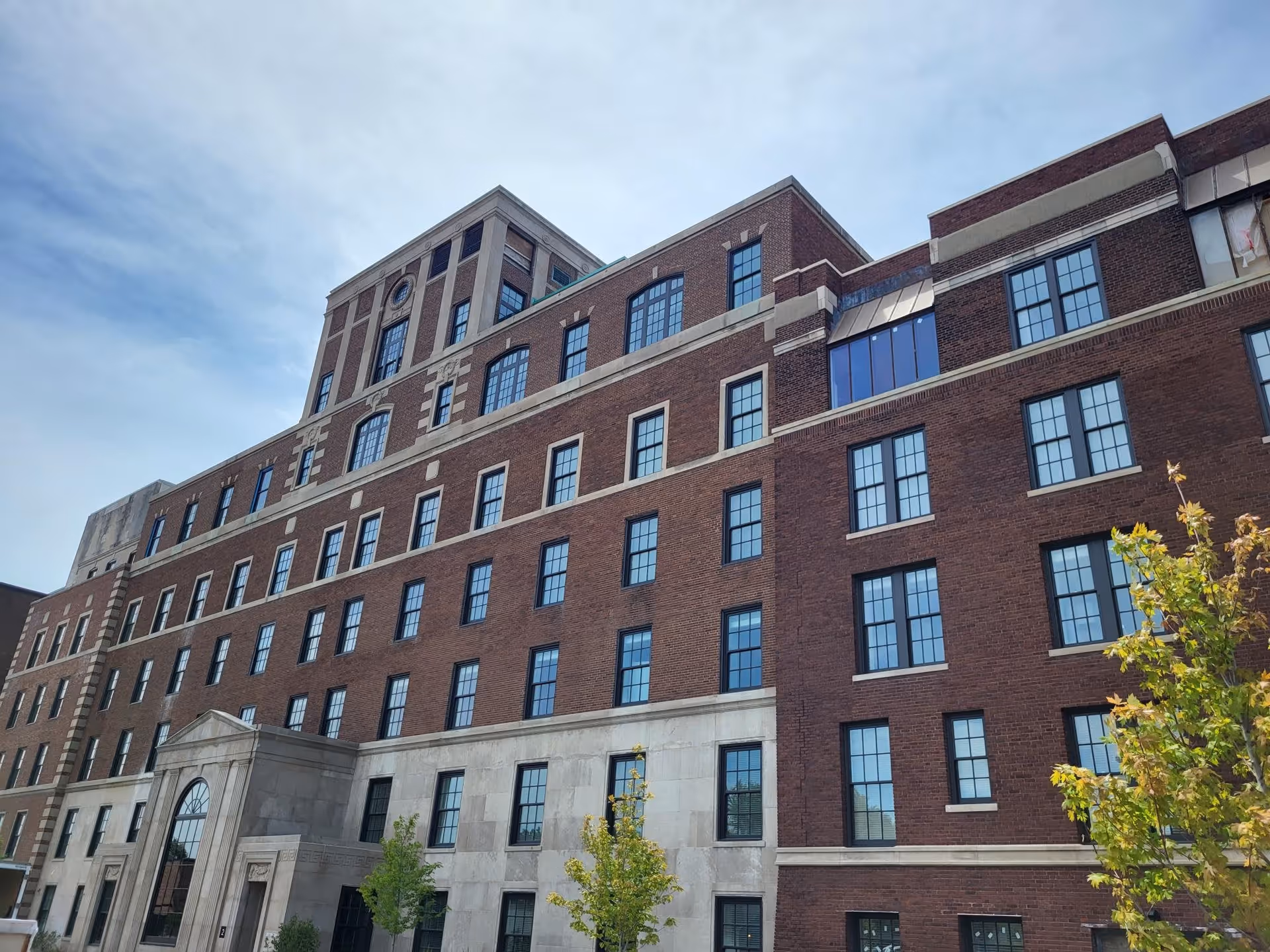 Multi-story red-brick building facade with many windows, a stone entrance, and trees in front under a blue sky.