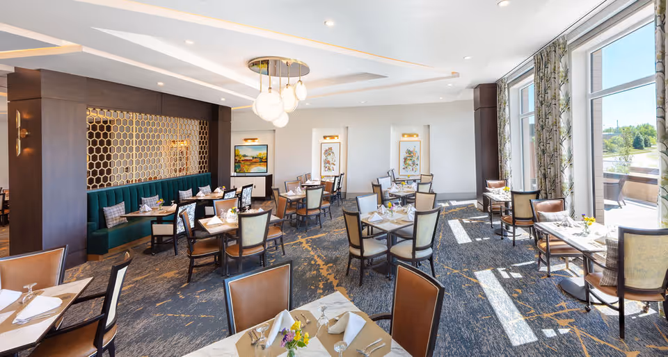 Bright and spacious dining room with multiple tables set with white napkins, glassware, and small flower arrangements. The room features large windows with patterned curtains allowing natural light to fill the space, a green upholstered bench along one wall, decorative wall art, and modern pendant lighting hanging from the ceiling.
