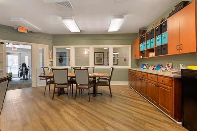 A well-lit room with wooden flooring featuring a rectangular table surrounded by six chairs. On the right side, there is a long wooden cabinet with multiple drawers and colorful storage bins on top. The walls are painted light green with white trim, and there are three windows with white frames on the far wall. The room appears clean and organized with some artwork and decorations on the cabinet. In the background, an open doorway leads to another area with more seating and large windows.