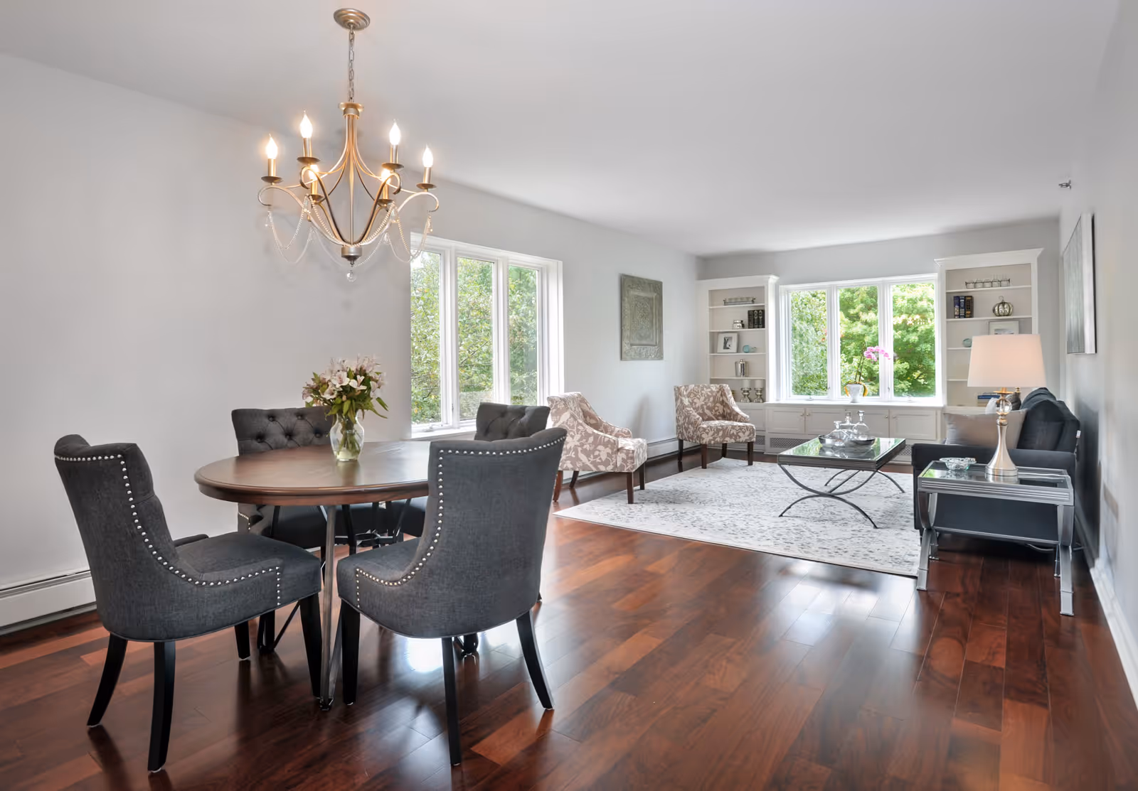 Open-plan living and dining area with a round wooden table and gray upholstered chairs, hardwood floors, and a seating area by large windows.