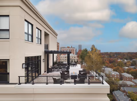 Outdoor terrace area of a senior living facility with seating arrangements including chairs and tables, overlooking a cityscape with trees and buildings under a partly cloudy sky.