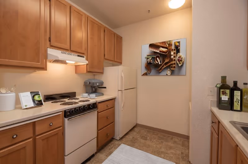 A compact kitchen with wooden cabinets, a white refrigerator, an electric stove with four burners, a coffee maker, and various kitchen items on the countertops. A framed picture of bread and olives hangs on the wall.