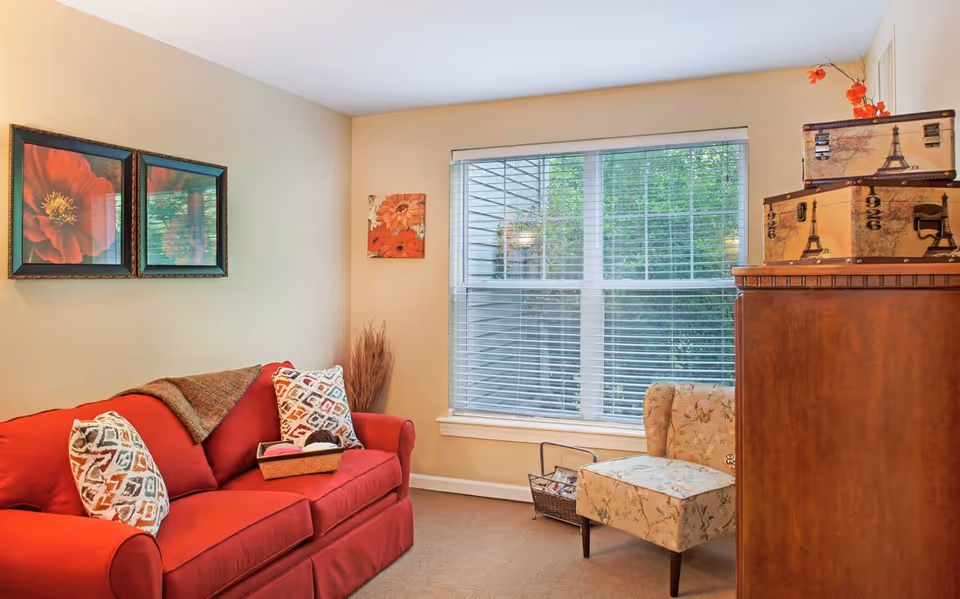 Cozy living room with a red sofa, patterned throw pillows, a floral accent chair, large window with blinds, wall art and a wooden cabinet.