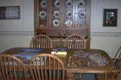 Wooden dining table with chairs, placemats and scattered puzzle pieces in front of a china hutch displaying plates.