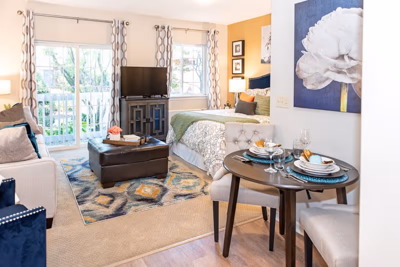 A bright and cozy studio apartment featuring a bed with patterned bedding, a small round dining table set for two, a comfortable sofa, a dark ottoman, a TV on a stand, and large windows with patterned curtains letting in natural light.