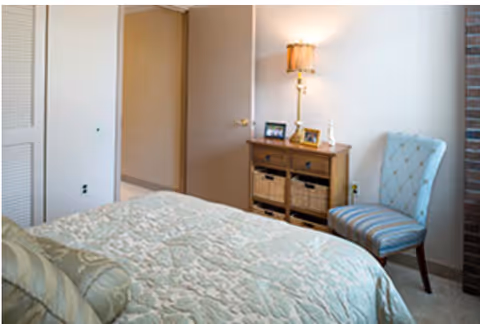 A bedroom with a bed covered in a light-colored patterned quilt and a matching pillow. There is a wooden dresser with baskets and framed photos on top, next to a lit table lamp. A blue upholstered chair with a tufted back is positioned near the dresser. The room has light-colored walls and an open door leading to another area.