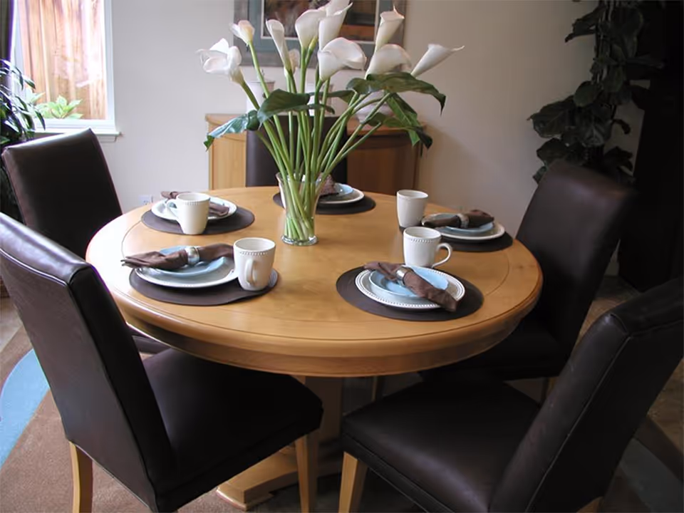 Round wooden dining table set for four with brown leather chairs and a vase of white calla lilies in the center.