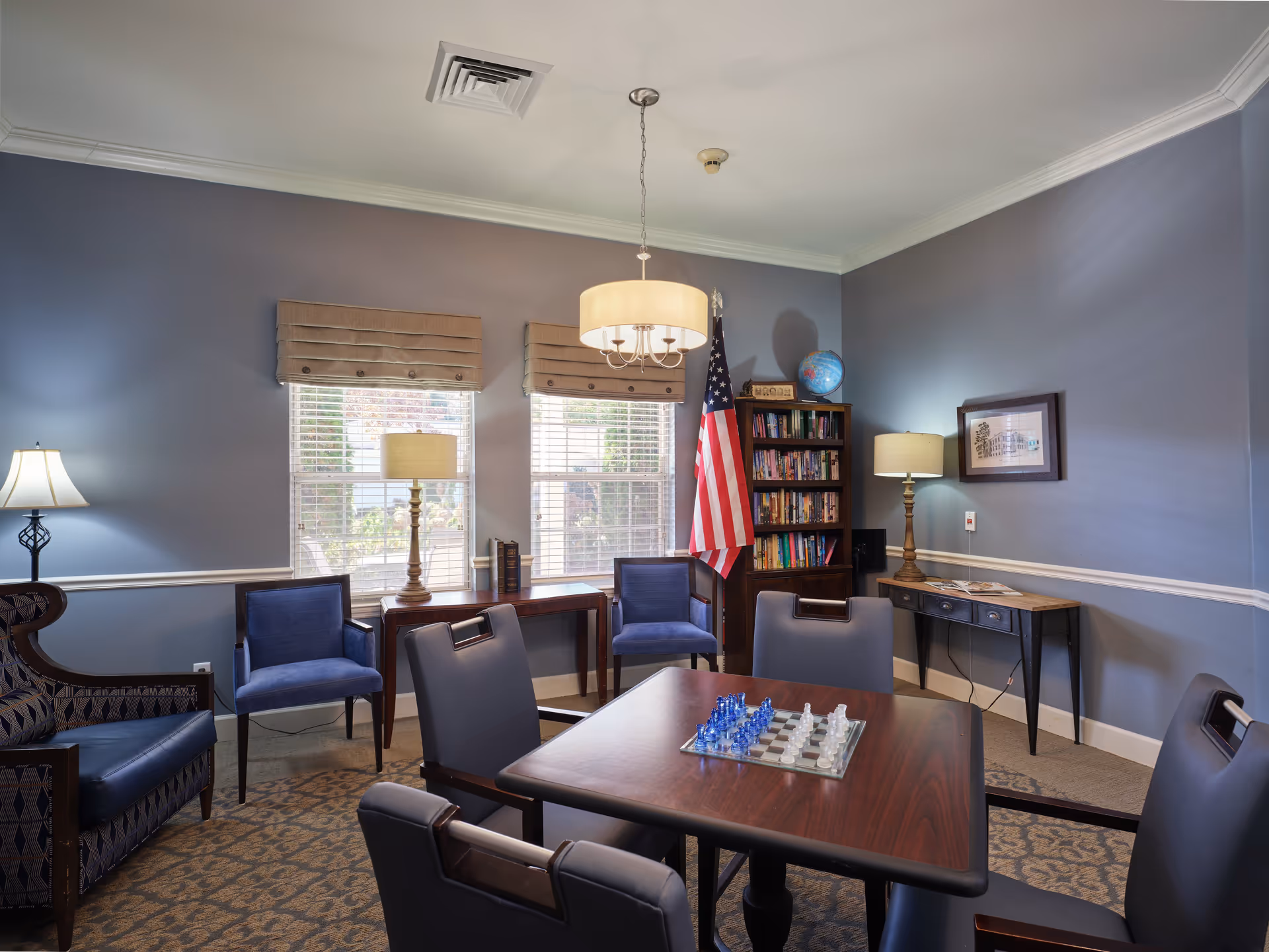 A cozy room with blue-gray walls featuring a square wooden table with a chess set on it, surrounded by four padded chairs. There are two blue armchairs and a patterned armchair along the walls. Two windows with beige Roman shades let in natural light. A bookshelf filled with books and a globe stands in the corner next to an American flag. Two table lamps and a ceiling light fixture illuminate the room. A framed picture hangs on one wall, and a small console table with magazines is against another wall.