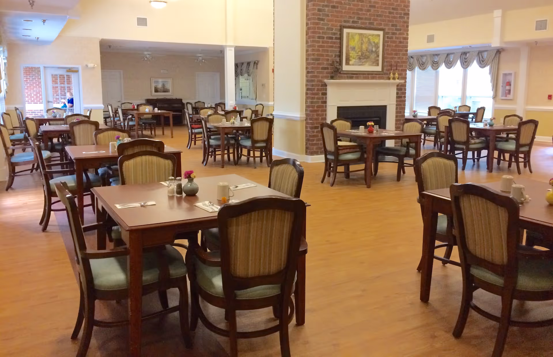 A spacious dining room in an assisted living facility with multiple wooden tables and chairs arranged neatly. Each table has place settings with utensils and small flower vases. There is a brick fireplace with a framed painting above it, and large windows with valances letting in natural light.