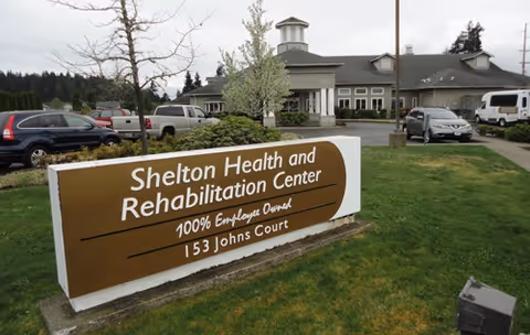 Exterior view of Shelton Health and Rehabilitation Center showing the building with a parking lot and several parked vehicles. In the foreground, there is a large brown and white sign with the facility's name, 'Shelton Health and Rehabilitation Center,' along with the text '100% Employee Owned' and the address '153 Johns Court.'