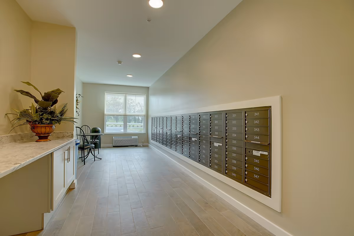 A bright hallway with a row of mailboxes mounted on the right wall, a window at the end letting in natural light, a small table with two chairs near the window, and a countertop with a potted plant on the left side.