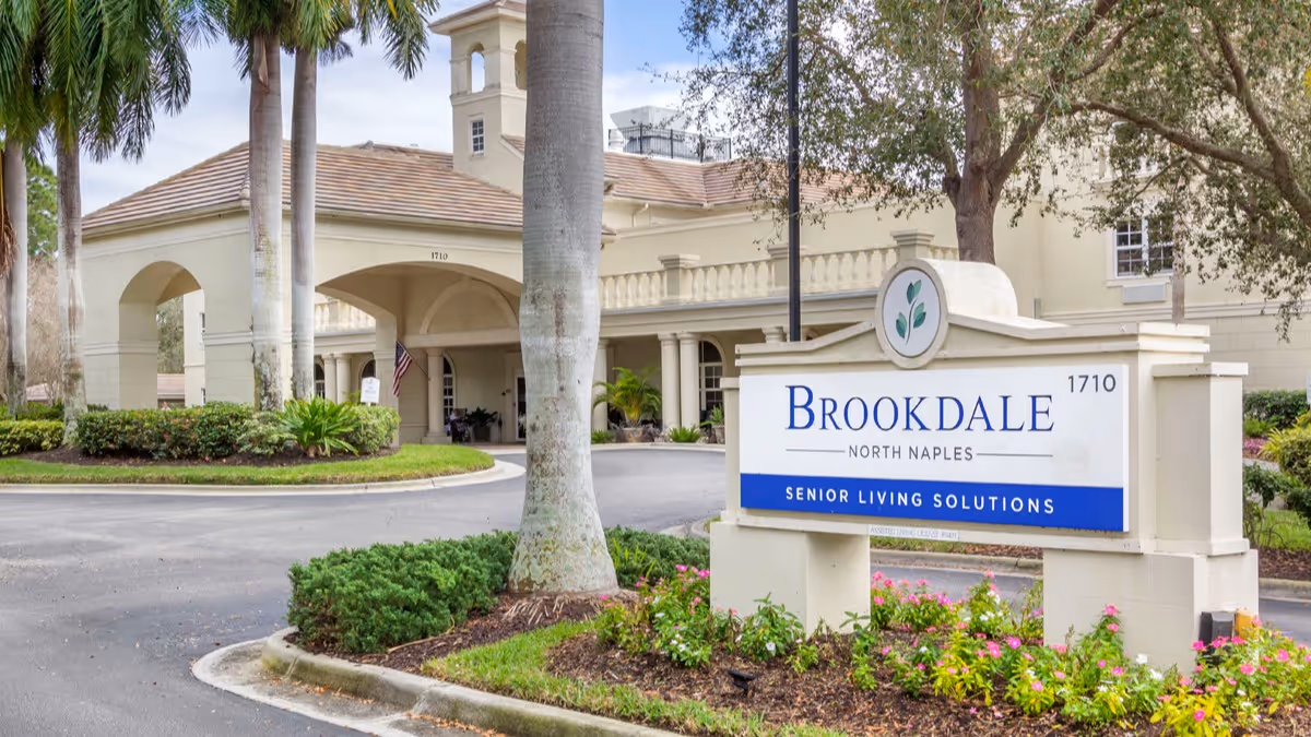 Front entrance of Brookdale North Naples senior living facility with a driveway, palm trees, and a large sign reading 'Brookdale North Naples Senior Living Solutions'.