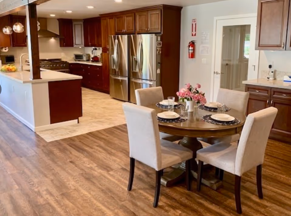 A modern kitchen and dining area with wooden flooring and cabinetry. The kitchen features stainless steel appliances including a double-door refrigerator and a stove with a range hood. The dining area has a round wooden table set with four beige upholstered chairs, plates, glasses, and a vase of pink flowers. The room is well-lit with recessed ceiling lights and pendant lights above the kitchen counter.