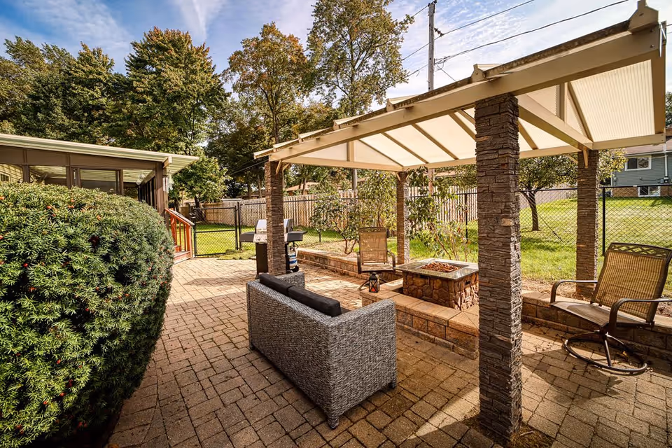 Outdoor patio area with a covered pergola supported by stone pillars, featuring a fire pit, a gray cushioned sofa, two chairs, and a grill. The patio is paved with bricks and surrounded by greenery and trees, with a fenced yard in the background.