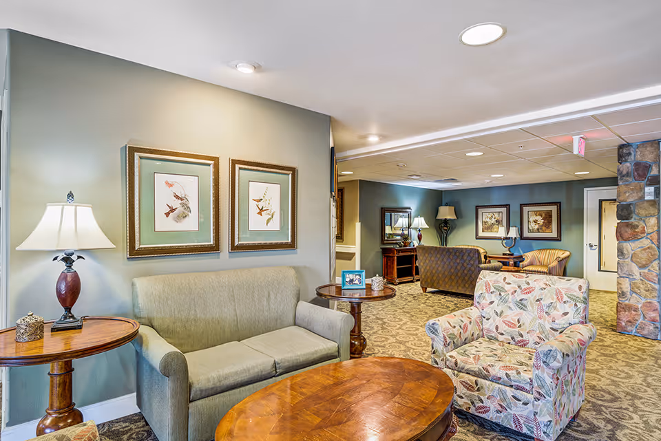 A cozy living room area in a senior living facility with a green sofa, a floral patterned armchair, wooden coffee and side tables, table lamps, framed bird artwork on the wall, and a carpeted floor. The room has soft lighting and a stone accent wall near a door in the background.